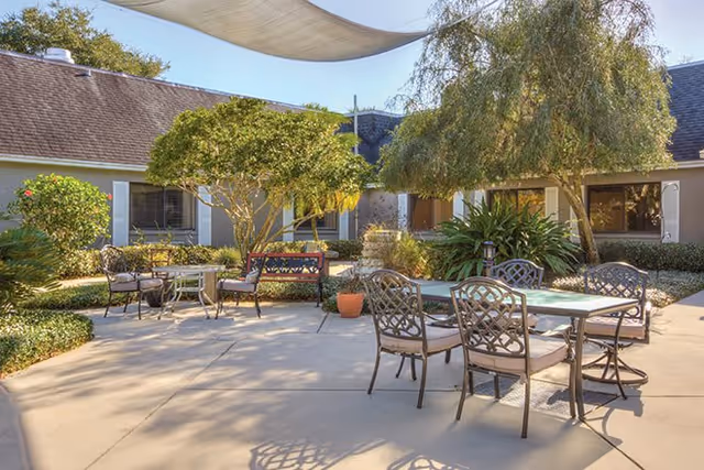 Outdoor courtyard area at Regency Oaks Health Center with several metal tables and chairs arranged on a concrete patio, surrounded by trees, shrubs, and a building with windows. A shade sail is stretched overhead providing partial shade.