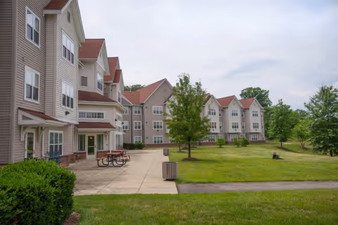 Exterior view of a senior living facility with multiple three-story buildings featuring beige siding and red roofs. There is a concrete patio area with picnic tables and benches, surrounded by well-maintained green lawns and trees under a cloudy sky.