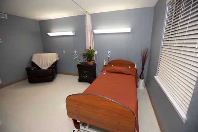 A simple room with a single hospital-style bed covered with a rust-colored blanket, a small black nightstand with a potted plant, a brown recliner chair with a beige throw draped over it, and a window with closed blinds. The walls are painted gray and the floor is light-colored.