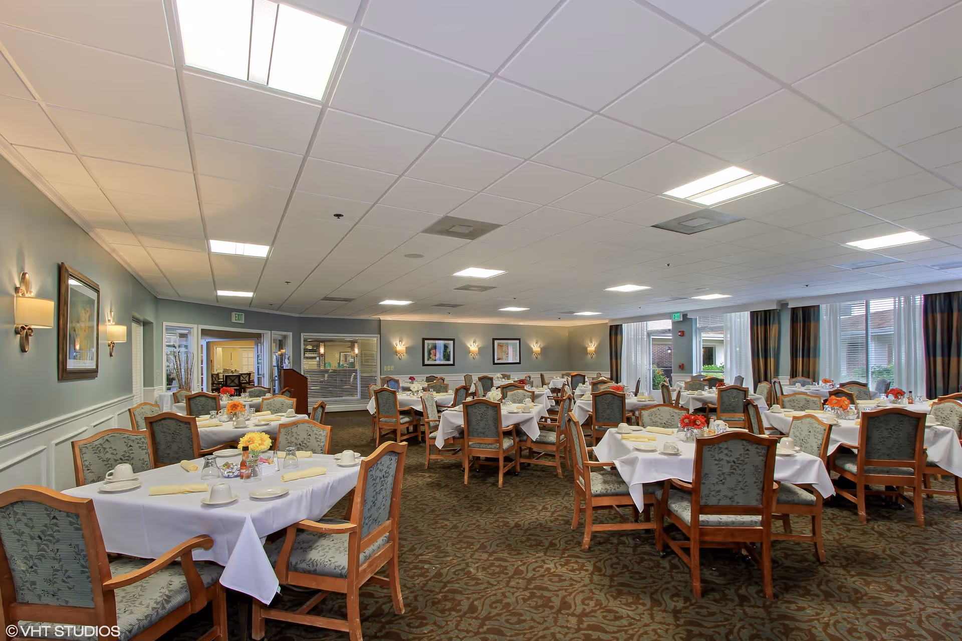 A spacious dining room with multiple tables covered in white tablecloths, each set with cups, plates, and napkins. The room has upholstered wooden chairs, patterned carpet, soft wall lighting, and large windows with curtains allowing natural light to enter.
