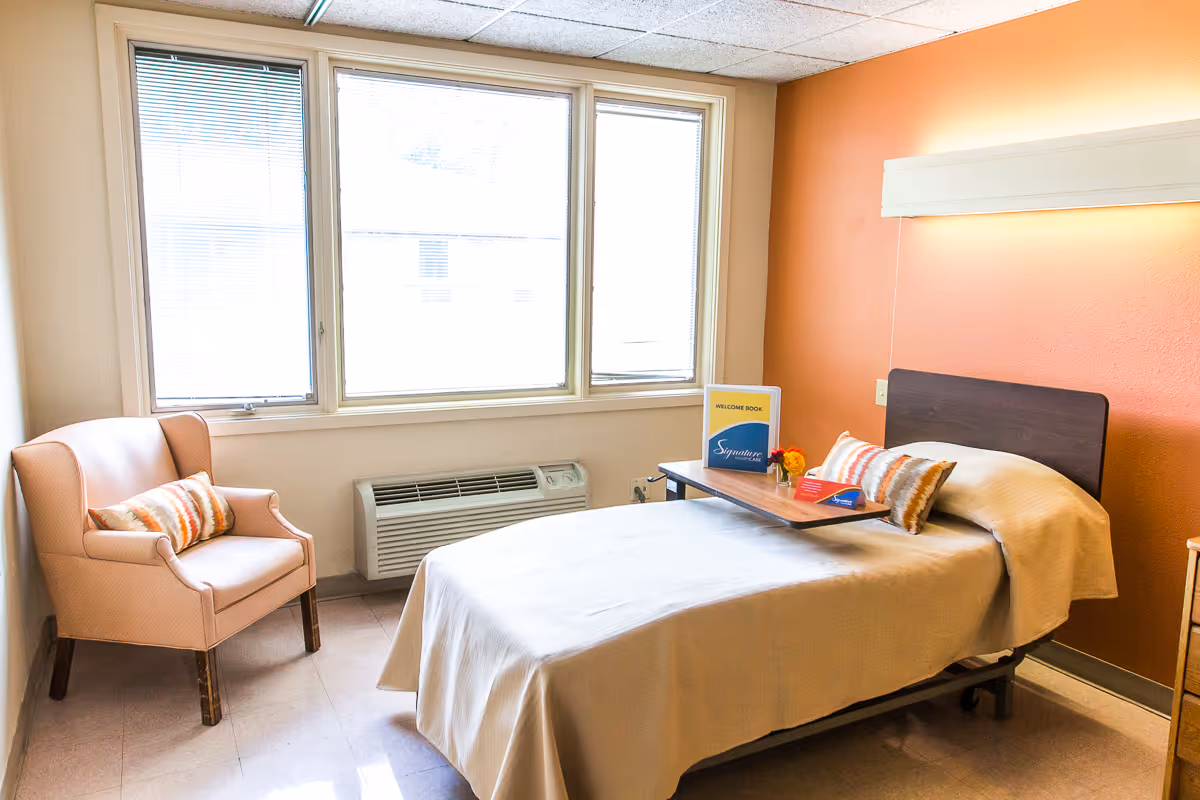 A tidy single-occupancy resident bedroom with a bed, armchair, large windows, and a welcome book on an overbed tray.