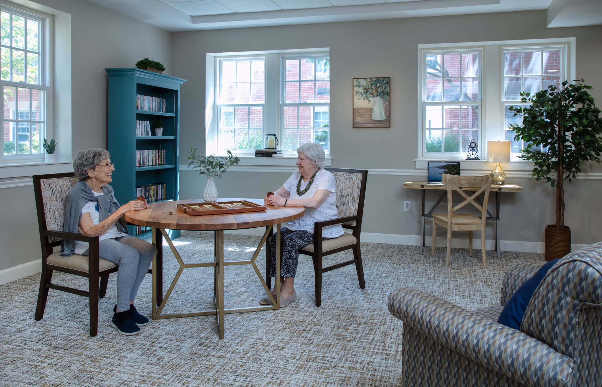 Two elderly women sitting at a round wooden table playing a board game in a bright, cozy room with large windows. The room has a patterned carpet, a blue bookshelf filled with DVDs, a small desk with a chair and a laptop, a potted plant, and a patterned armchair in the foreground.