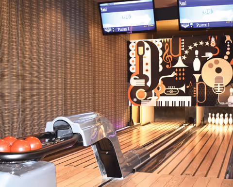 Indoor bowling alley with bowling balls on a rack, a polished wooden lane, pins set up at the end, and two screens displaying scores above the lane. The back wall features a colorful mural with musical instruments and notes.