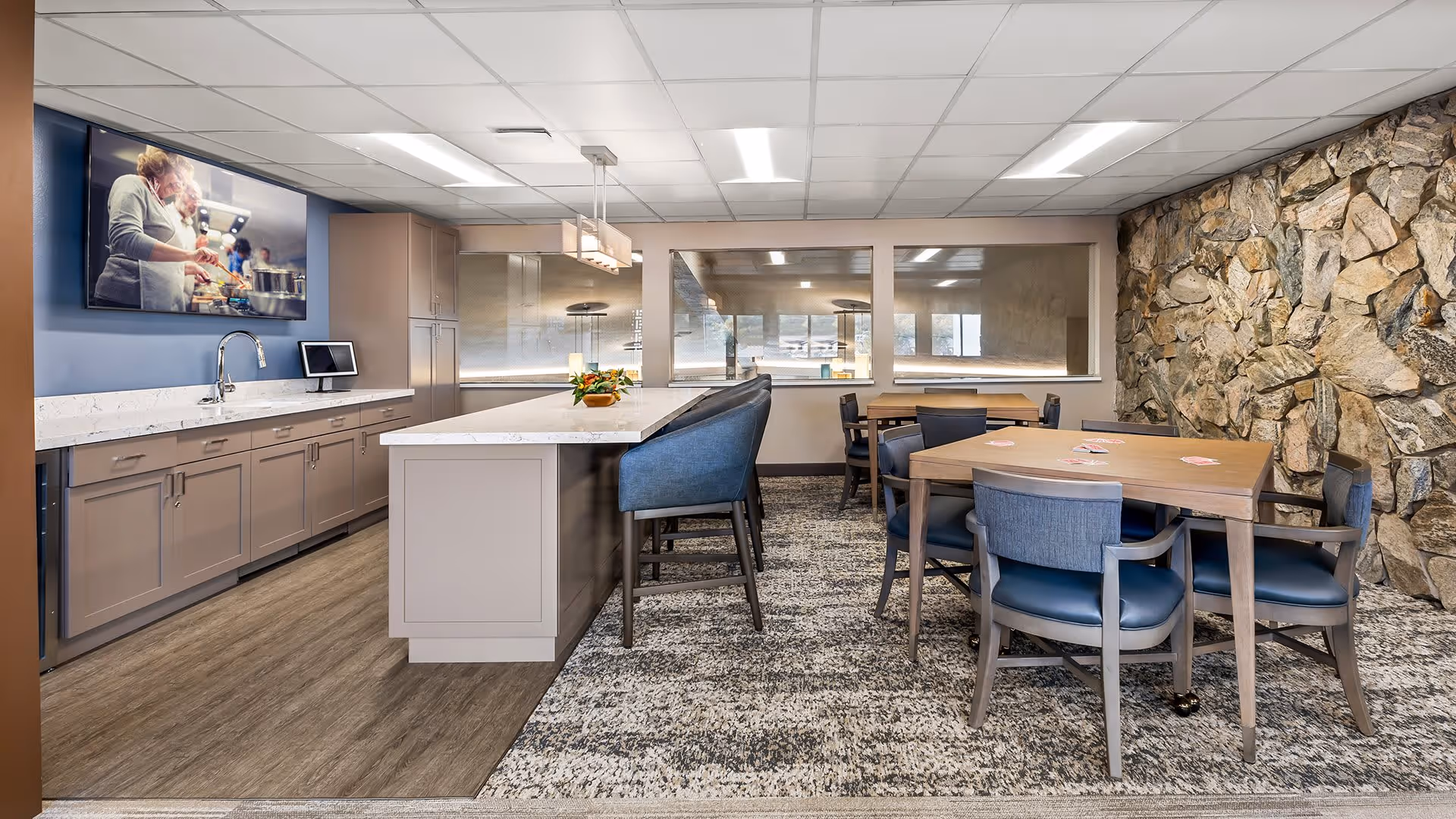 A modern communal dining and kitchen area with a kitchen island featuring a white countertop and blue cushioned bar stools. The kitchen has beige cabinets, a sink, and a mounted TV showing a cooking scene. There are several wooden tables with blue cushioned chairs on a patterned carpet, and a stone accent wall on the right side.