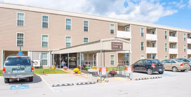 Front entrance of a three-story senior living building with a covered drop-off, parking spaces, and a sign reading 'Friendship House'.