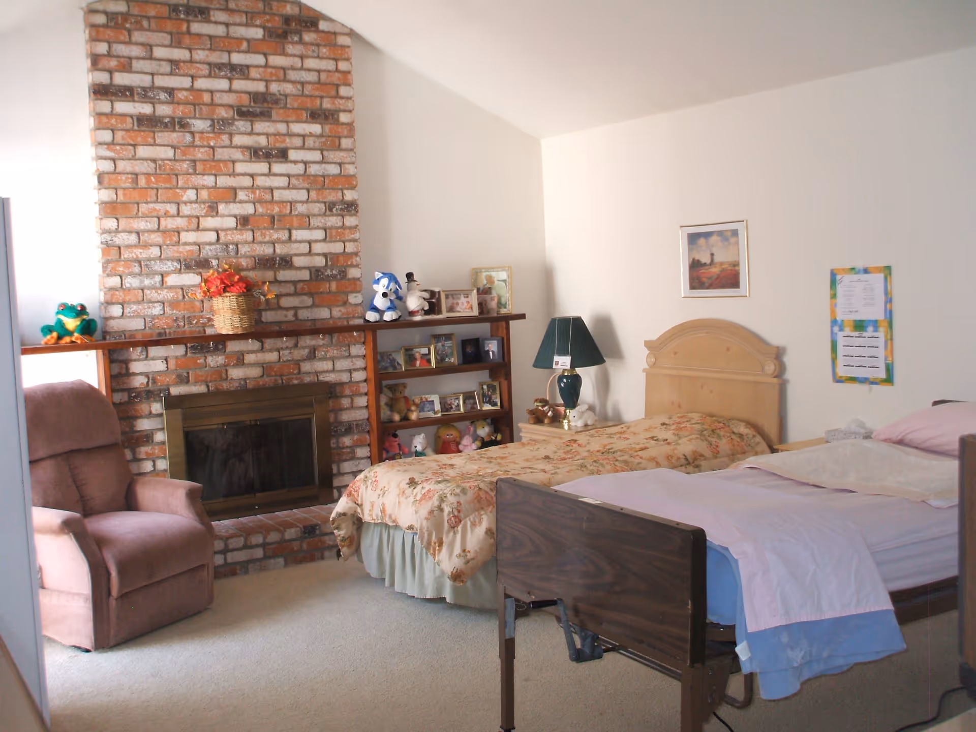 A furnished bedroom with two beds, a recliner, a brick fireplace, shelving with photos and stuffed animals, and a bedside lamp.