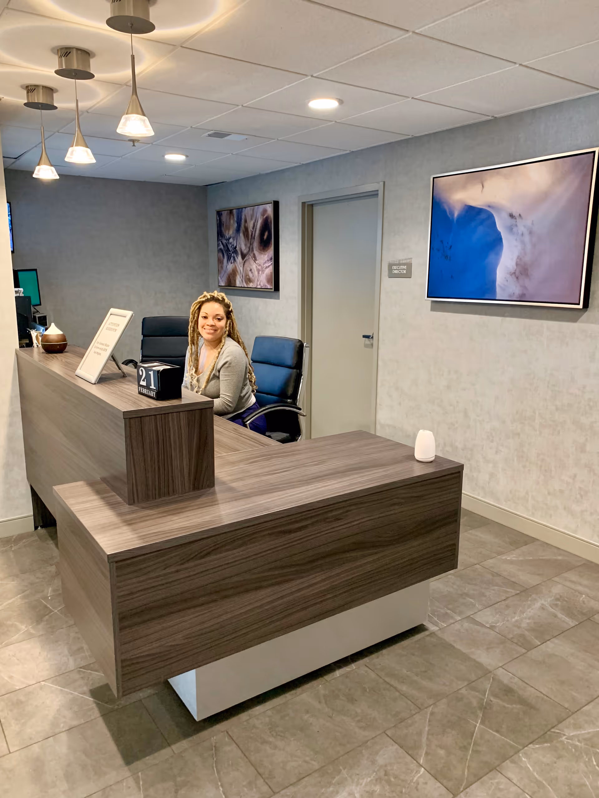 A woman with long braided hair sitting behind a modern wooden reception desk in a well-lit office area. The walls are light gray with two abstract framed artworks hanging. The floor is tiled, and there are three pendant lights hanging from the ceiling above the desk.
