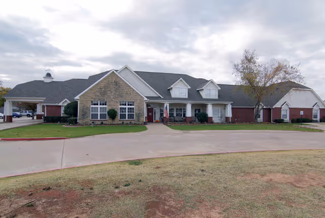Front exterior view of a single-story senior living facility building with a combination of brick and stone facade, multiple windows, a covered entrance with white columns, an American flag near the entrance, and a driveway in front. The sky is cloudy and there is a tree on the right side of the building.