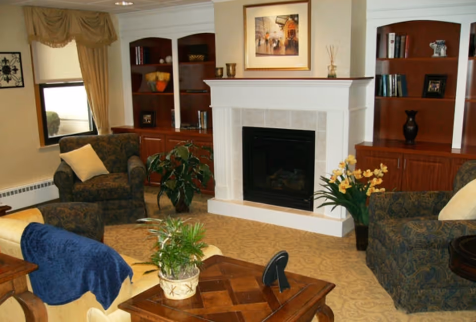 Cozy common-area living room with a central fireplace, built-in shelving, upholstered chairs, plants, and a coffee table.