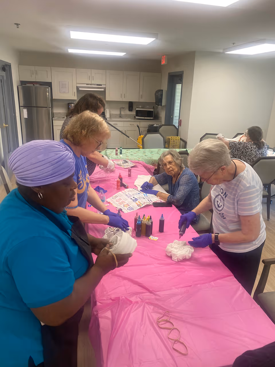 A group of seniors and caregivers gathered around a pink-covered table doing tie-dye crafts in a communal room with a kitchenette in the background.