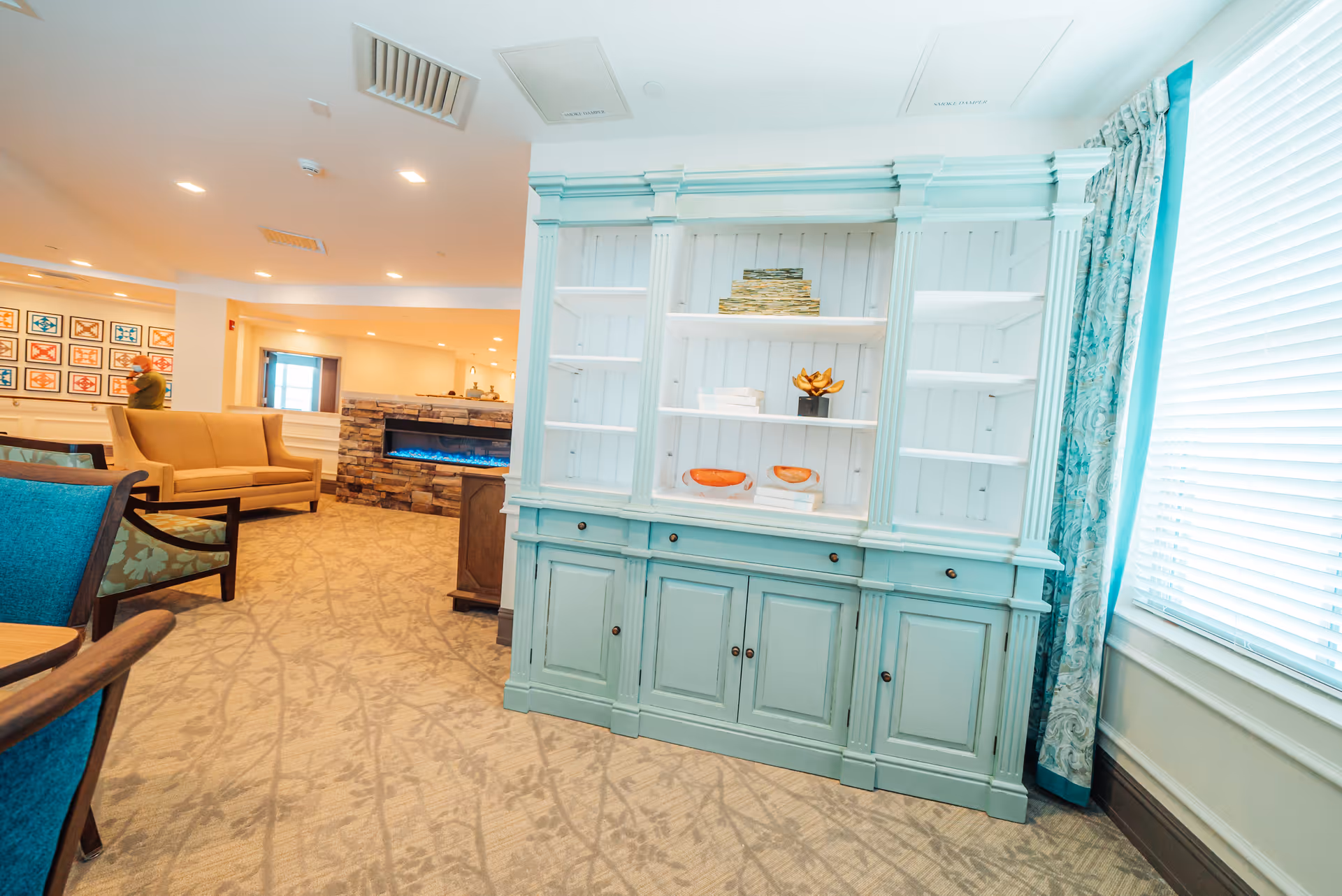 A bright and spacious living area in a senior living facility featuring a light blue wooden cabinet with shelves displaying decorative items, a beige sofa, blue upholstered chairs, a stone fireplace with a blue flame, and large windows with patterned curtains allowing natural light to fill the room.