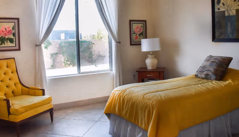 Bright bedroom featuring yellow bedspread, armchair, window, and nightstand in a senior facility.