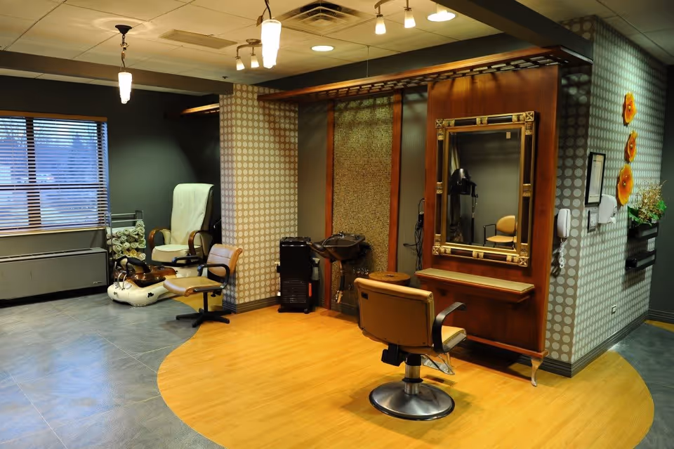 Interior of a salon area in a senior living facility with a styling chair facing a large mirror mounted on a wooden panel. To the left, there is a pedicure station with a white foot bath and a white reclining chair. The walls have a patterned wallpaper, and there are decorative orange flowers on the right wall. The floor is a combination of wood and tile.
