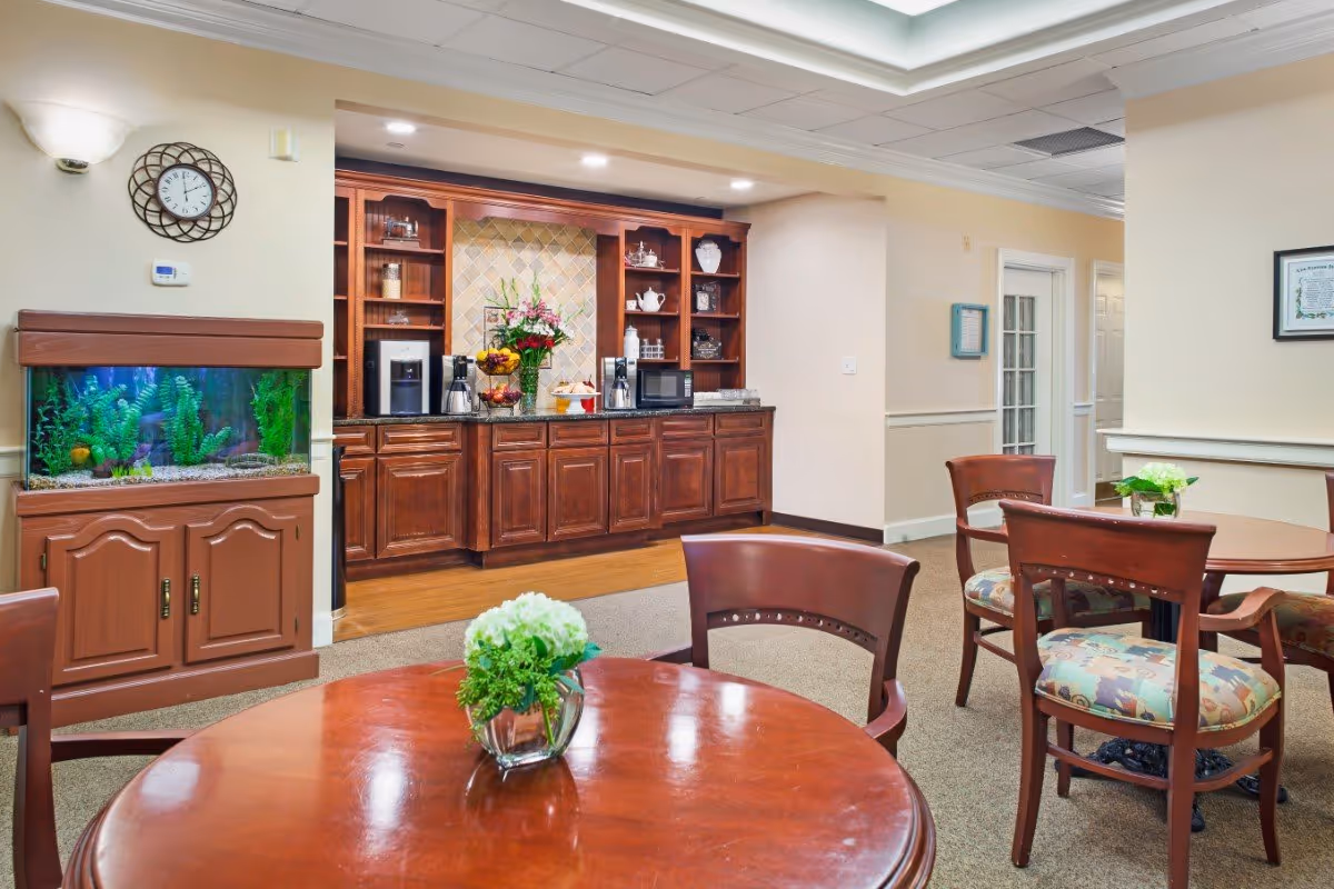 A bright communal dining/lounge area with round wooden tables and chairs, a fish tank, and a built-in kitchenette and coffee station.