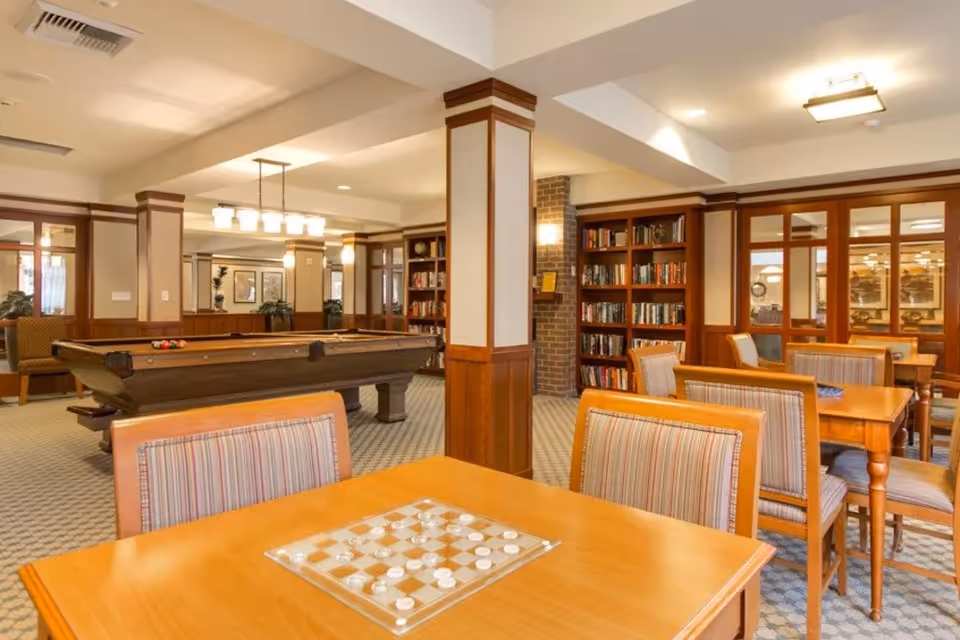 A cozy common room with wooden tables and striped cushioned chairs, a checkers game set on one table, a pool table in the background, and bookshelves filled with books along the walls. The room features warm lighting and wood-paneled columns.