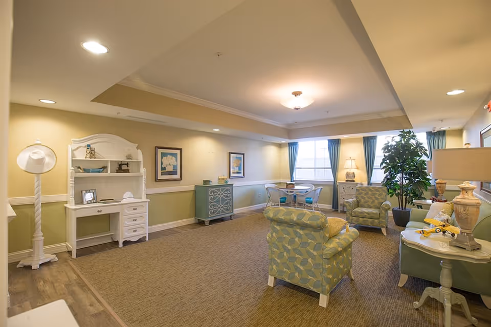 A cozy living room area in a senior living facility with patterned armchairs, a green sofa, a small round table with chairs near windows with blue curtains, a white desk with shelves, framed artwork on the walls, a tall coat rack with a white hat, and decorative lamps and plants providing a warm ambiance.