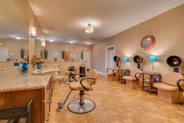Interior view of a salon area in a senior living facility with multiple salon chairs, hair drying stations, a large mirror, and decorative plants and flowers. The room has warm lighting and a clock on the wall.
