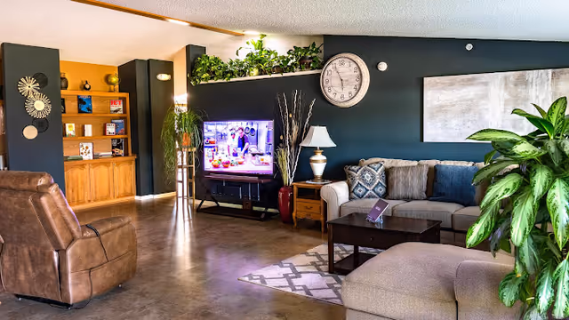 A cozy living room area with a brown leather recliner, a beige sectional sofa with patterned cushions, a dark wooden coffee table, and a TV showing a cooking show. The walls are painted dark green with a large clock and abstract artwork hanging. There are several green plants and a wooden bookshelf with decorative items in the background.