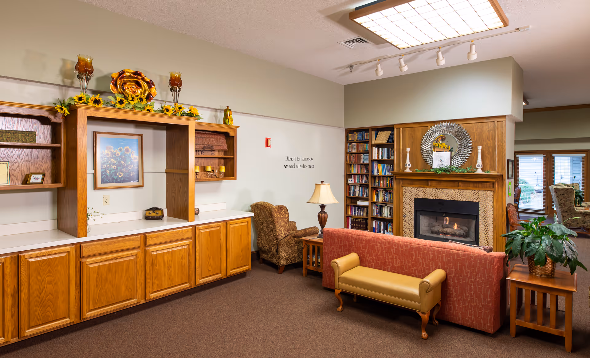 A cozy living room area in an assisted living facility featuring a fireplace with a decorative mirror above it, bookshelves filled with books, a red sofa, a yellow bench, an armchair, a side table with a lamp, and wooden cabinetry with decorative items and a framed sunflower picture. The room has carpeted floors and a ceiling light fixture.