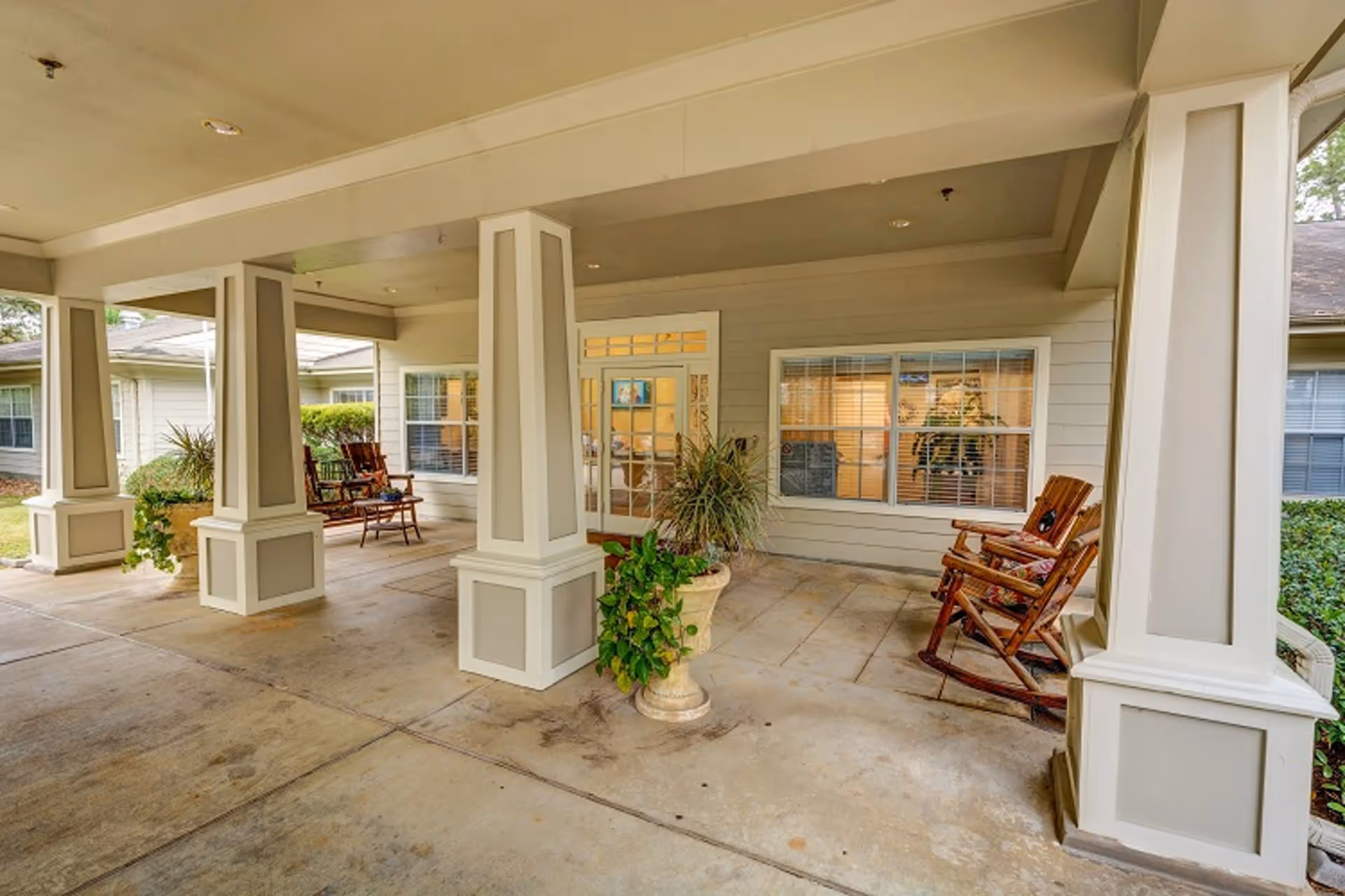 Covered outdoor patio area with large square columns, wooden rocking chairs with cushions, potted plants, and windows looking into the building interior.
