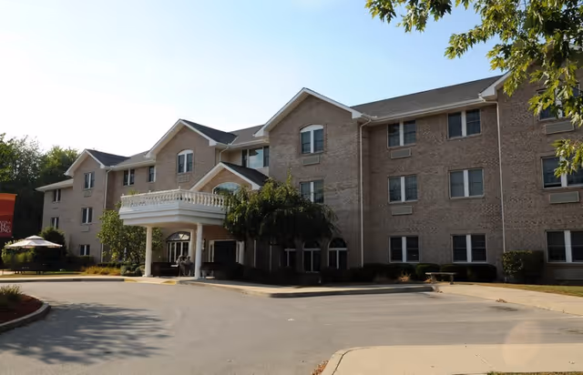 Exterior view of a three-story brick senior living facility with a covered entrance and a driveway in front. Trees and landscaping surround the building under a clear sky.