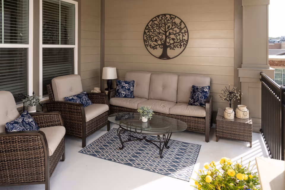 A cozy outdoor patio area with a wicker sofa and two matching armchairs, all with beige cushions and blue patterned throw pillows. A glass-top coffee table sits on a blue and white patterned rug. There is a side table with decorative items and a lamp, and a metal wall art piece of a tree hangs on the beige paneled wall. Yellow flowers are visible in the foreground, and a black railing borders the patio.