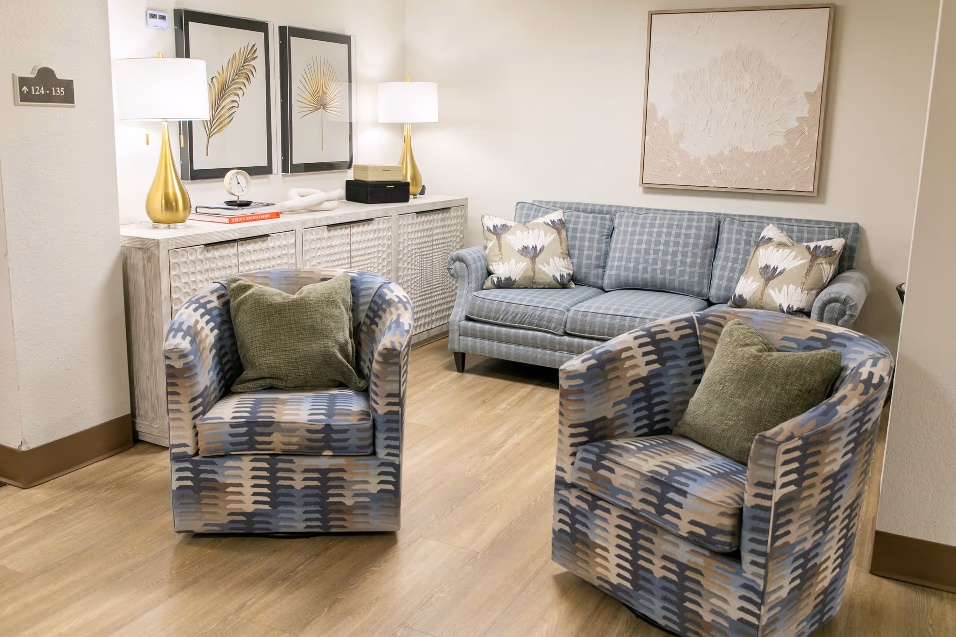 A cozy sitting area in a senior living facility featuring two patterned armchairs with green cushions, a blue plaid sofa with floral pillows, a white textured sideboard with two gold lamps, decorative items, and framed botanical artwork on the walls.