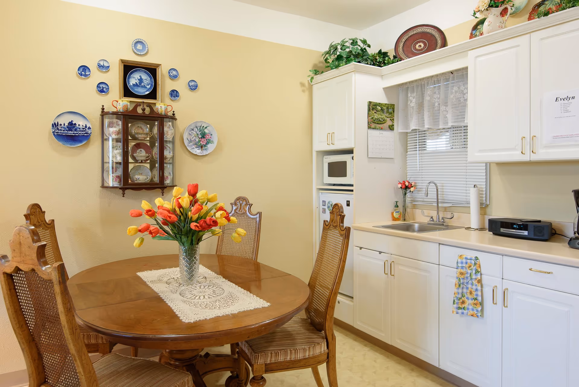 A cozy kitchen and dining area with a round wooden table and four chairs. A vase with red and yellow tulips sits on a lace table runner on the table. The kitchen has white cabinets, a sink under a window with lace curtains, a microwave, and a small refrigerator. Decorative plates and a small cabinet with china are mounted on a beige wall. There are plants and decorative items on top of the cabinets.