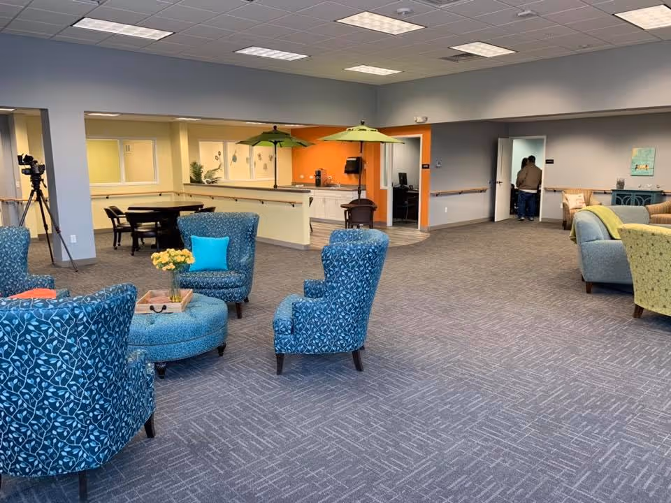 A spacious senior living facility common area with blue patterned armchairs arranged around a round ottoman with a tray and yellow flowers. In the background, there is a small dining area with tables and chairs, a kitchenette with two green umbrellas over small tables, and a hallway with two people standing near an open door. The walls are painted in neutral tones with an orange accent wall in the kitchenette area.