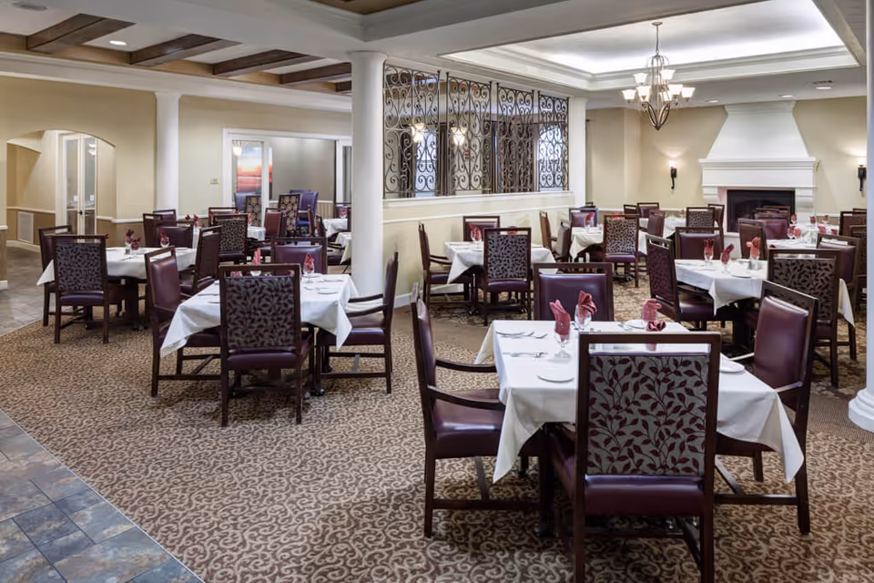 A spacious dining room with multiple tables covered in white tablecloths, each set with folded red napkins, glasses, and silverware. The room features patterned carpet, beige walls, white columns, a decorative wrought iron partition, and a white fireplace. The ceiling has recessed lighting and a chandelier.