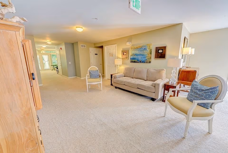 Carpeted communal lounge with a sofa, accent chairs, lamps, artwork, and a hallway leading further into the facility.