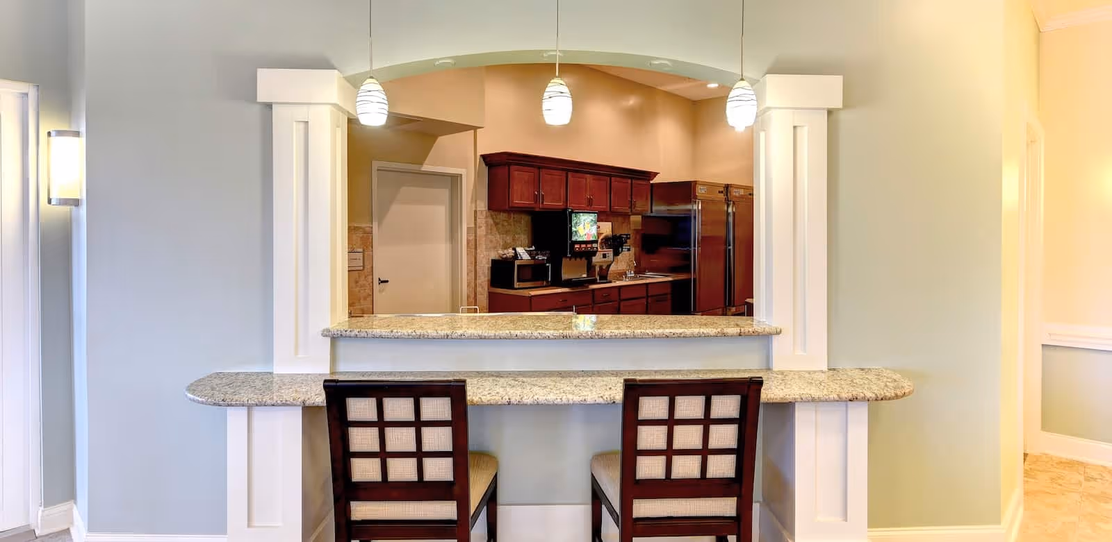Interior view of a senior living facility kitchen area seen through a large pass-through window with a granite countertop and two wooden chairs with cushioned seats. The kitchen features dark wood cabinets, a stainless steel refrigerator, a microwave, and a coffee machine. Three pendant lights hang above the countertop, and the walls are painted in soft neutral tones.