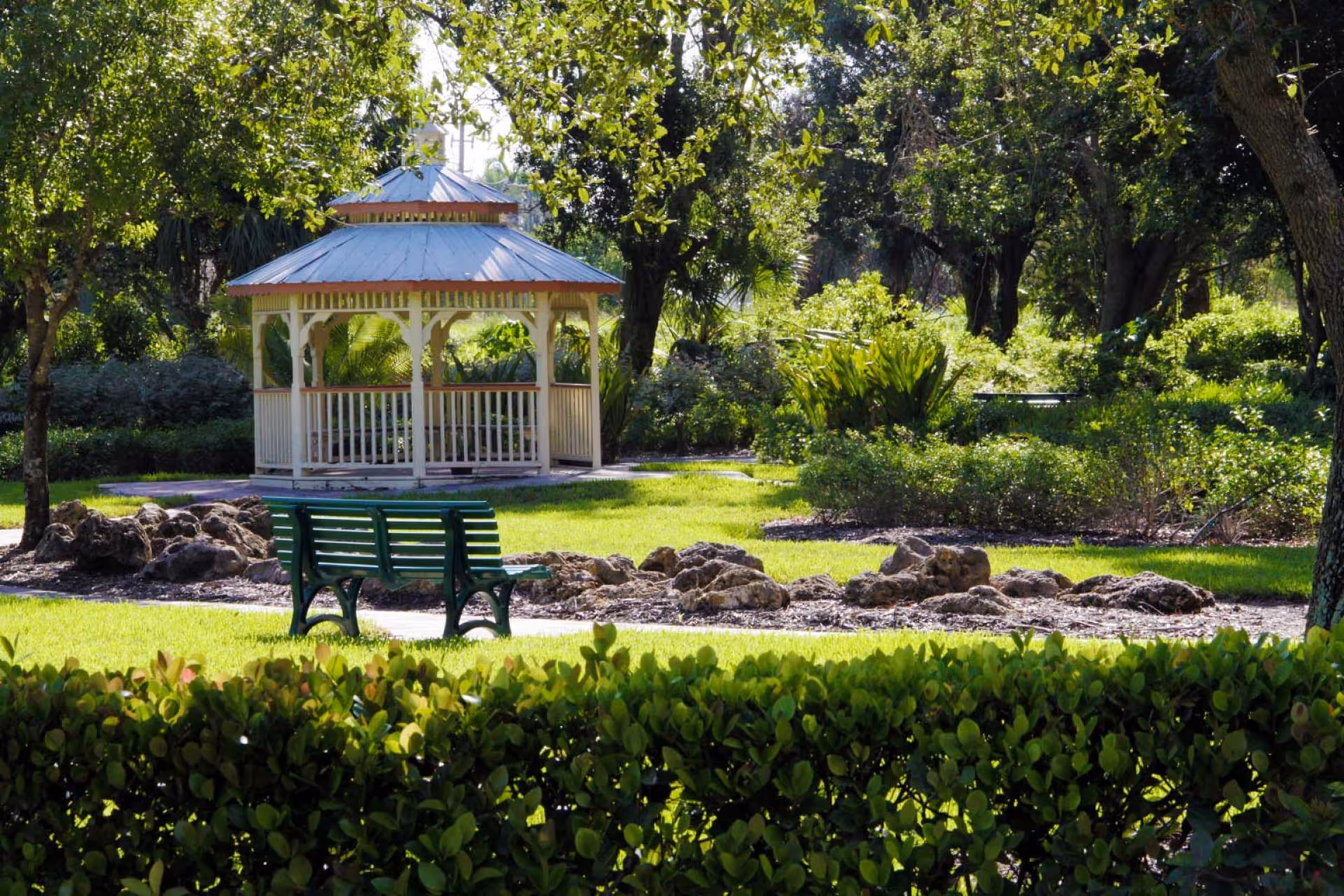 A white gazebo sits in a sunlit landscaped garden with a green park bench and leafy trees.