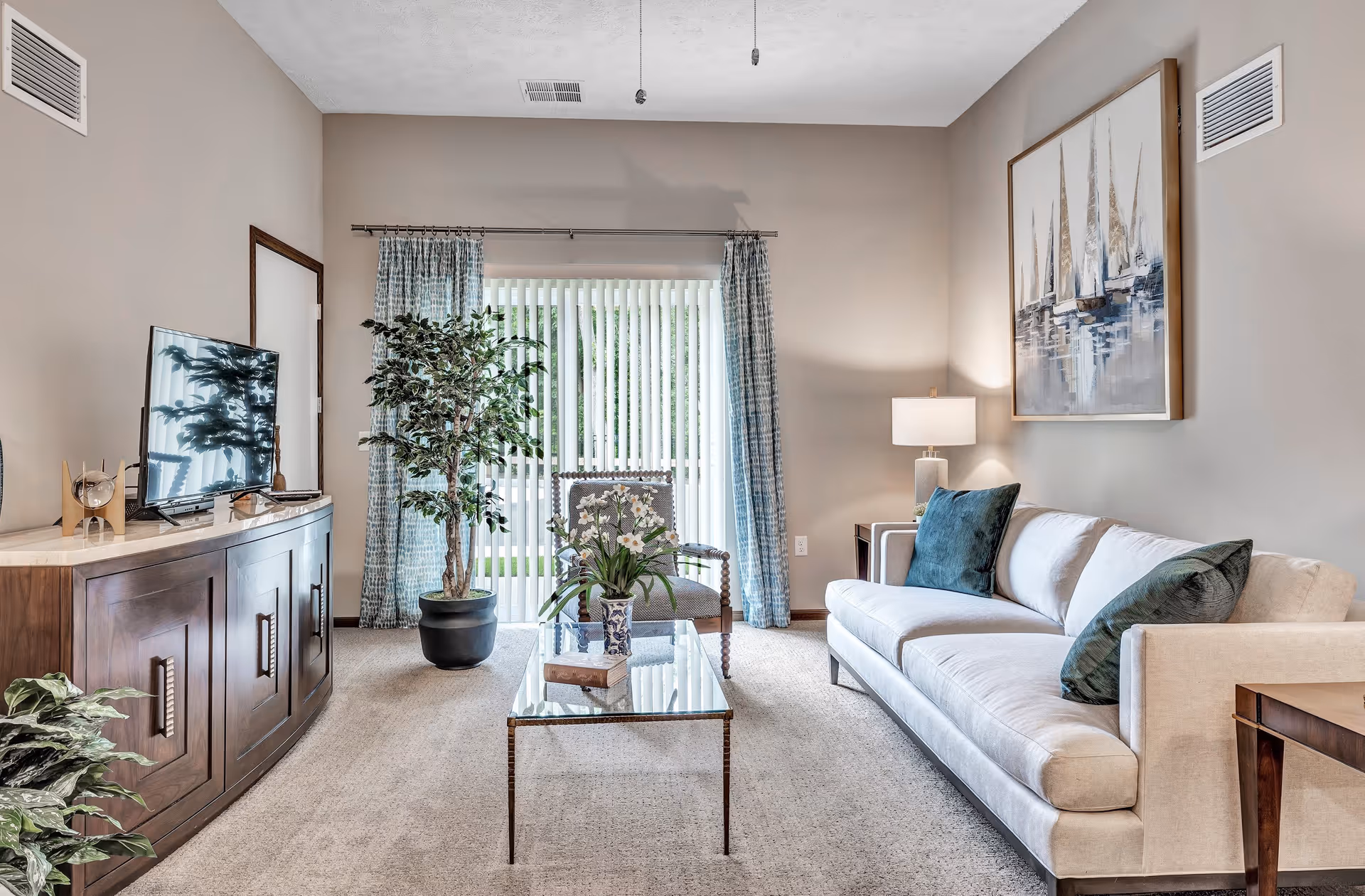 A bright and cozy living room in Greenway Square Retirement Community featuring a beige sofa with dark green pillows, a glass coffee table with a flower vase and a book, a wooden sideboard with a flat-screen TV, a patterned armchair, potted plants, blue patterned curtains, and a large painting of sailboats on the wall.