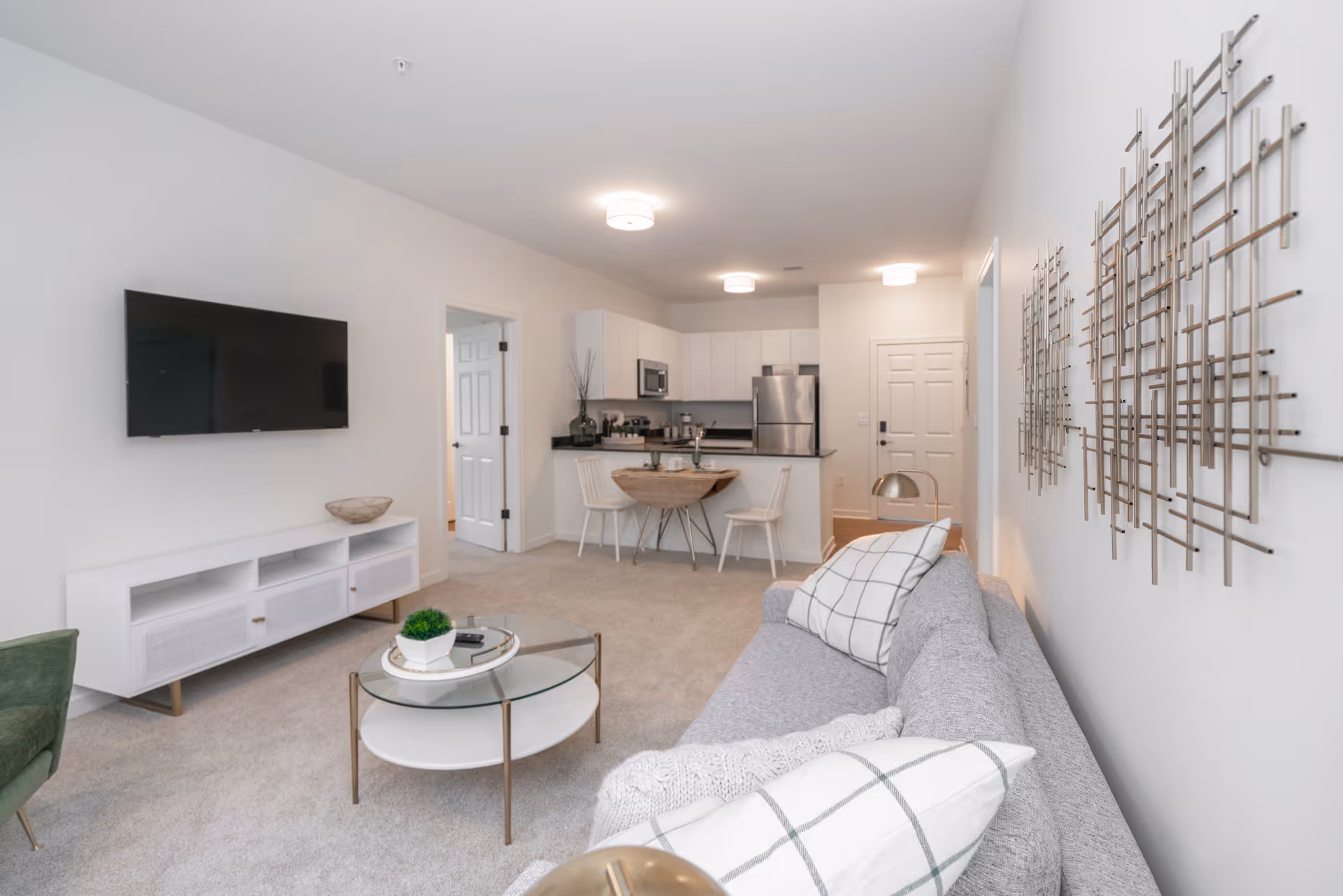 A modern living room and kitchen area in an apartment. The living room features a gray sofa with white and gray checkered pillows, a round glass coffee table with a small plant and remote controls, and a white TV stand with a mounted flat-screen TV. The kitchen has white cabinets, stainless steel appliances, a small round dining table with two white chairs, and a black countertop. The walls are white with decorative metal wall art on one side.