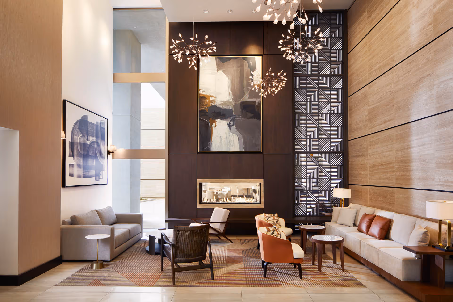 A modern and elegant living room area with high ceilings, featuring a large beige sofa with brown and beige cushions, two orange and white armchairs, a gray sofa, several small round wooden tables, and a contemporary fireplace set in a dark wood-paneled wall. Above the fireplace is an abstract painting, and to the right is a tall decorative panel with geometric patterns. The room is well-lit with natural light from large windows and stylish chandeliers hanging from the ceiling.