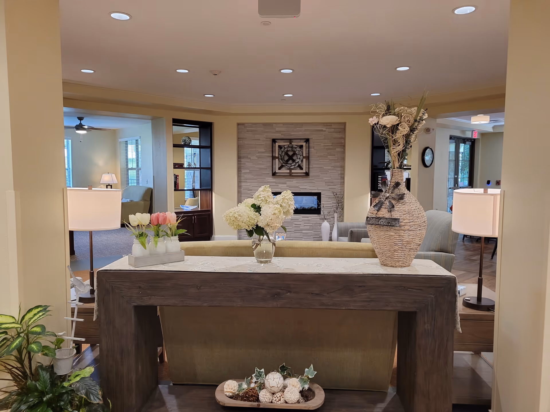 Bright, furnished common living area with a console table holding floral arrangements, sofas, lamps, and a fireplace in the background.