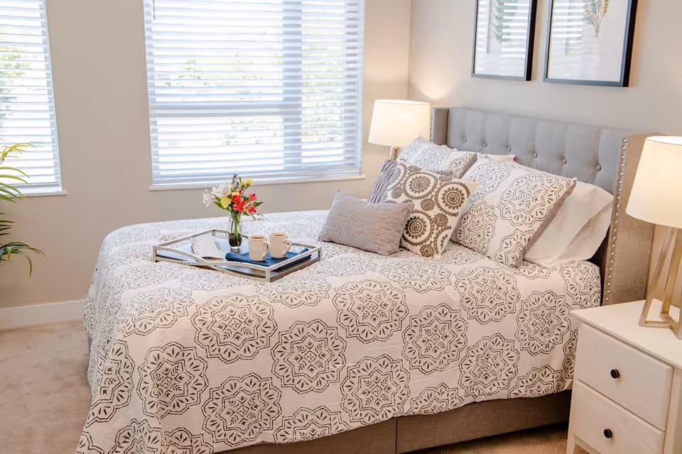 A neatly made bed with a patterned white and gray bedspread and multiple decorative pillows in a bright bedroom. There is a tray on the bed holding two coffee mugs, a book, and a small vase with flowers. Two bedside tables with lamps flank the bed, and two framed botanical prints hang on the wall above the headboard. A window with white blinds allows natural light into the room.