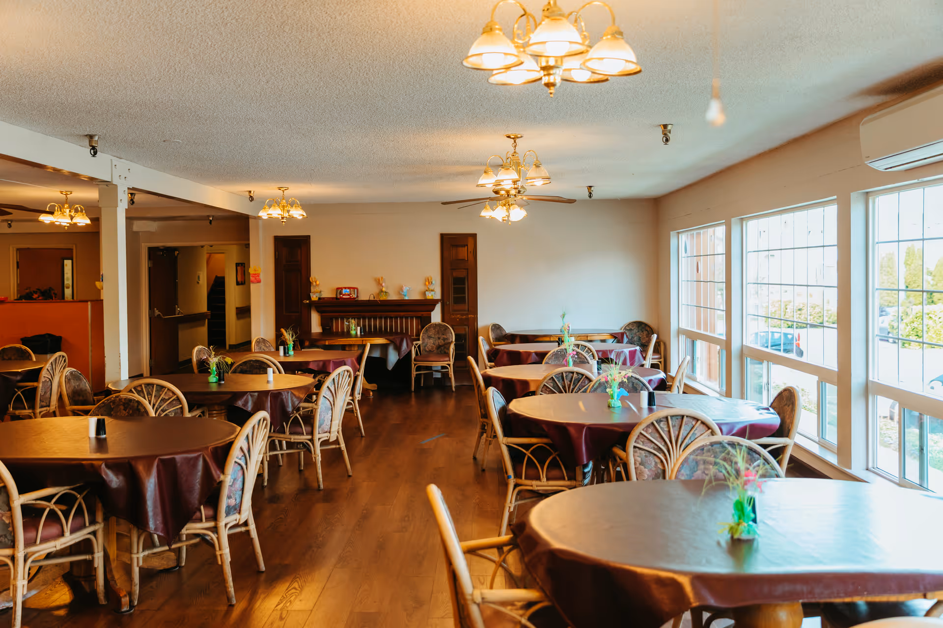 Dining room with round tables covered in tablecloths and wicker chairs, lit by ceiling fixtures and large windows along one side.