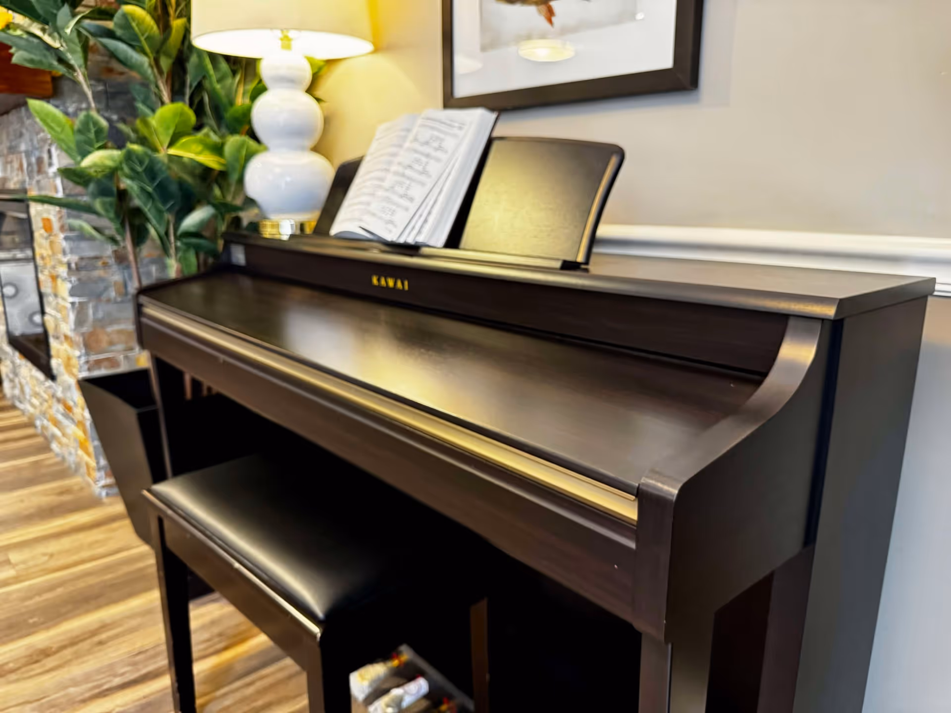 A dark wooden Kawai piano with a matching bench in front of it. On top of the piano is an open sheet music book and a white lamp with a round base. To the left, there is a green leafy plant and a stone wall with a fireplace. The floor is wooden, and a framed picture is partially visible on the wall behind the piano.