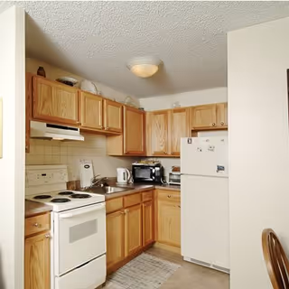 A compact kitchen with wooden cabinets, a white electric stove, a white refrigerator with magnets, a microwave, a toaster oven, a kettle, and a sink. There is a small rug on the floor and a wooden chair partially visible on the right side.