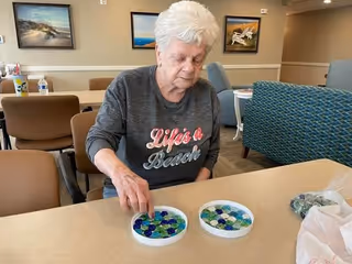 An elderly woman with white hair is sitting at a table in a common area, playing with colorful glass beads placed in two shallow white dishes. The room has beige walls with framed pictures and comfortable chairs in the background.