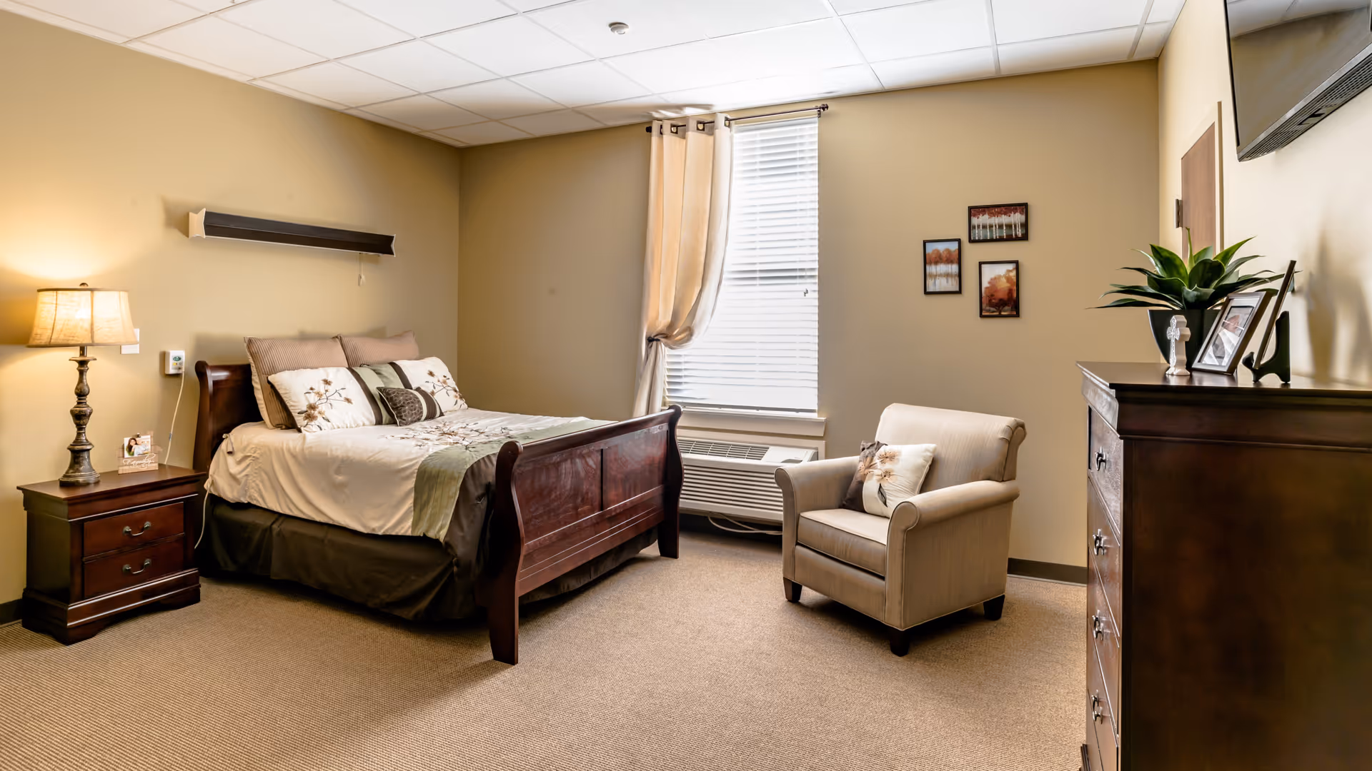 A cozy bedroom in a senior living facility with a wooden bed adorned with pillows and a floral bedspread, a nightstand with a lamp, a beige armchair with a decorative pillow, a window with beige curtains, a wooden dresser with a plant and framed photos, and three small framed pictures on the wall.