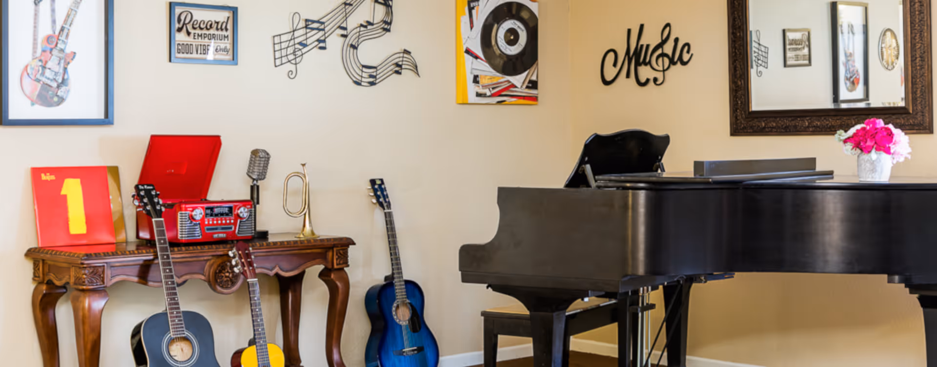A music-themed room with a black grand piano, a wooden table holding a red record player, a vintage microphone, a small trumpet, and three acoustic guitars leaning against the table and wall. The wall is decorated with framed pictures, musical notes, a vinyl record, and the word 'Music' in decorative script. A mirror and a vase with pink flowers are also visible on the piano.