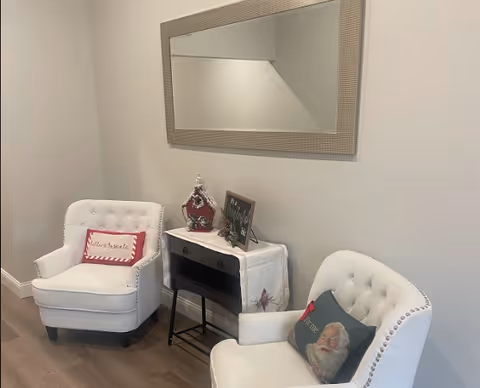 Two white tufted armchairs with decorative pillows flank a small table topped with decor beneath a large wall mirror in a seating area.