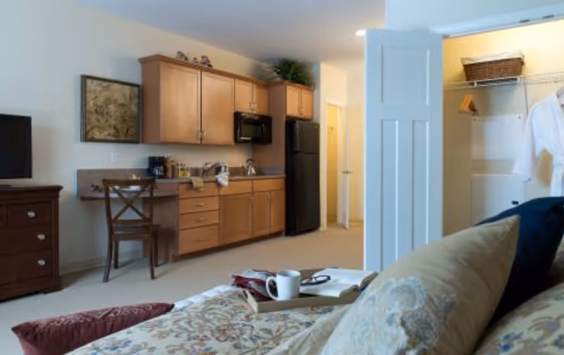 Interior view of a senior living facility room showing a small kitchenette with wooden cabinets, a black refrigerator, microwave, and coffee maker. In the foreground, there is a bed with patterned bedding and pillows, a tray with a cup and book on the bed. To the right, an open closet with a hanging white shirt and a basket on the shelf is visible. A wooden chair is placed near the kitchenette, and a TV is on a wooden dresser to the left.