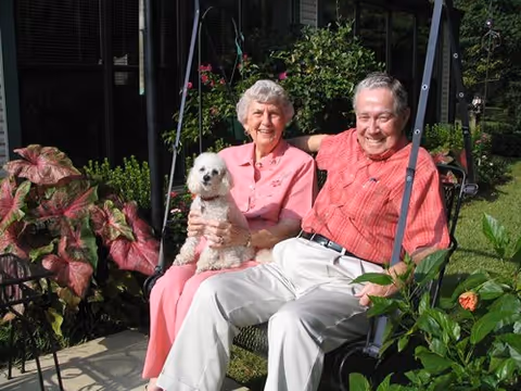 An elderly couple sitting together on an outdoor swing in a garden. The woman is holding a small white dog on her lap. Both are smiling and surrounded by green plants and flowers.