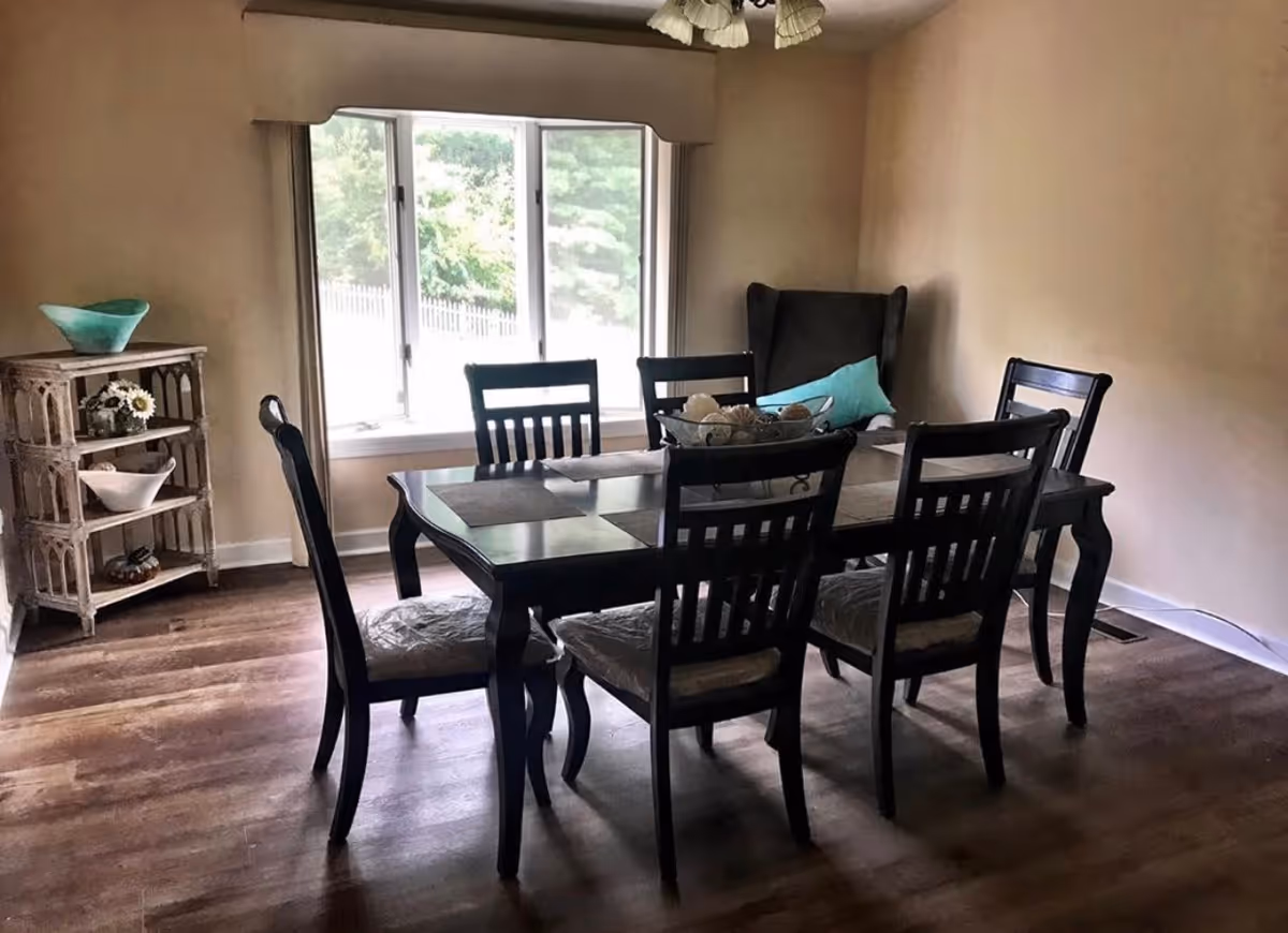 A dining room with a dark wooden table and six matching chairs. The table has placemats and a decorative bowl with assorted items. There is a large window with a view of greenery outside, a cushioned armchair with a blue pillow in the corner, and a small wooden shelf with decorative bowls and flowers on the left side. The room has wooden flooring and beige walls.