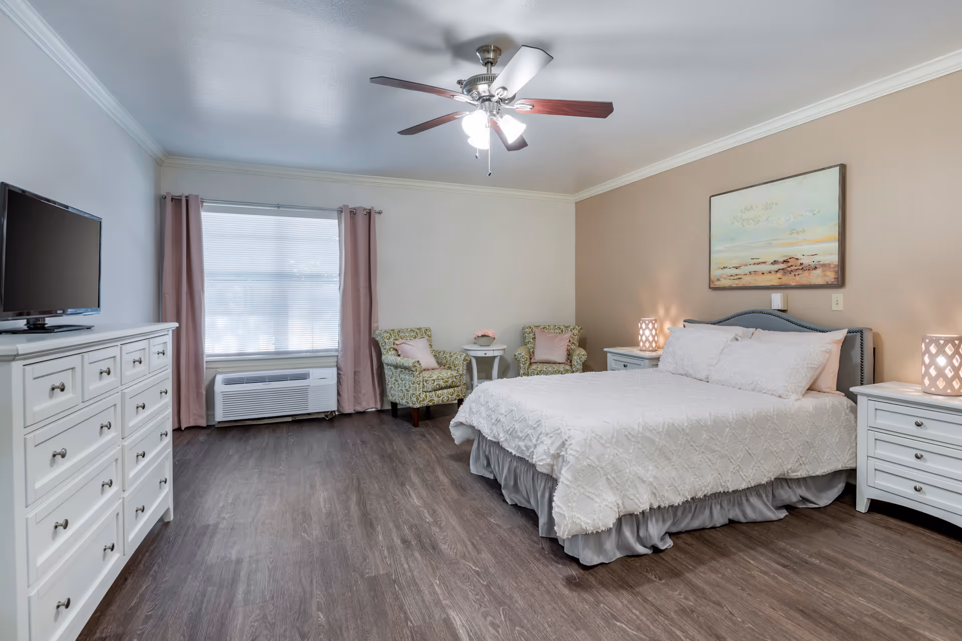 A well-lit bedroom with a large bed covered in white bedding, two white nightstands with lamps on either side, a dresser with a TV on top, two patterned armchairs with pink cushions, a small round table between the chairs, a window with pink curtains, and a ceiling fan with lights.