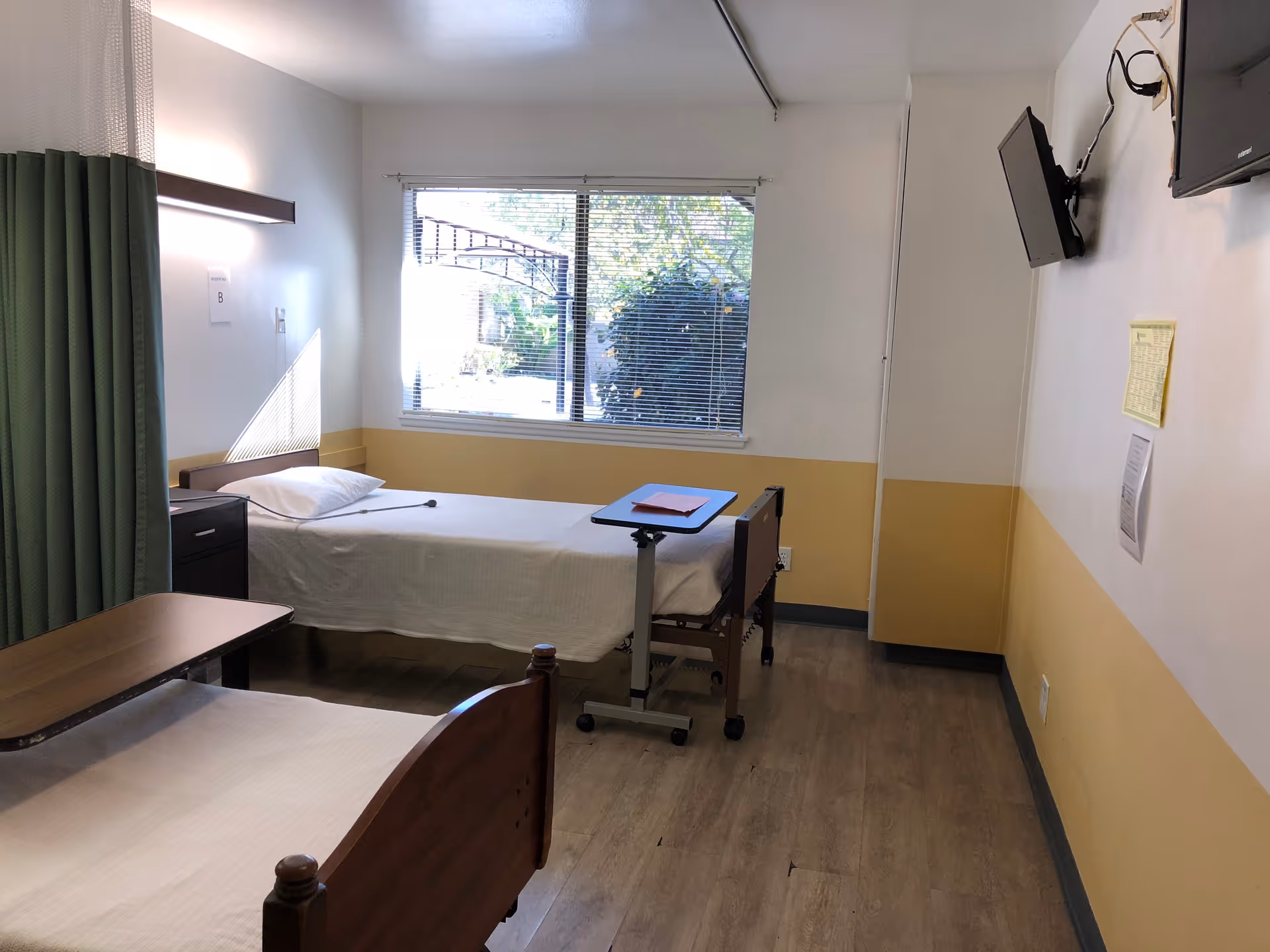 A simple, clean room with two single beds, each with white bedding and a pillow. One bed has an adjustable overbed table with a red folder on it. There is a window with blinds letting in natural light, a green privacy curtain partially visible on the left, and two wall-mounted televisions on the right wall. The walls are painted white with a yellow lower half, and the floor is wood.